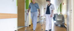 Ein älterer Patient an Krücken geht in Begleitung einer Physiotherapeutin einen Gang entlang